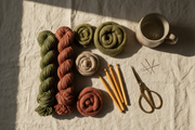 A warm, artistic flat lay featuring hand-dyed yarn skeins, wool roving, crochet hooks, scissors, embroidery needles, and a ceramic mug on a linen background. Natural morning light, soft shadows, neutral tones with muted greens, clay, and cream accents. Cozy, handmade, artisan craft aesthetic, photographed from above.