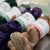 Several skeins of variegated yarn arranged in a close-up product photo. The yarn colors include deep purple, forest green, and a warm beige/tan blend, each with white paper bands around the center.