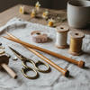 close-up of artisan fiber tools — wooden knitting needles, brass scissors, measuring tape, spools of thread — arranged artfully on linen cloth, warm neutral lighting, cottagecore studio aesthetic
